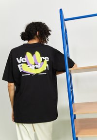 Zwarte t-shirt met een multikleuren smiley graphic en de tekst "Venture Berlin" op de achterkant. Korte mouwen, losse pasvorm, gemaakt van katoen.