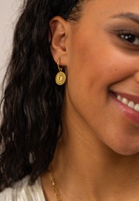 Goldene Creolenohrringe mit detaillierten, kreisförmigen Anhängern und komplizierten Mustern, die an einem strukturierten Ohrläppchen neben einem lächelnden Gesicht zur Schau gestellt werden.