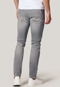 Mężczyzna w wąskich szarych dżinsach i białych trampkach stoi tyłem na jednolitym jasnym tle.