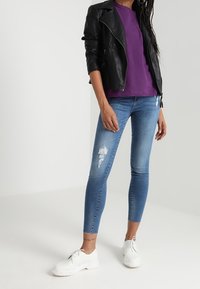 Černá kožená motorkářská bunda přes fialový top, doplněná světle modrými úzkými džíny s prošoupáním a bílými šněrovacími botami.