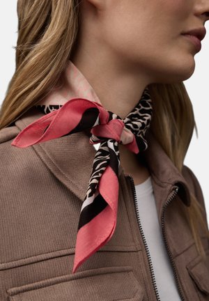 Femme portant une veste beige et une écharpe à motifs rose, noir et crème nouée en nœud papillon autour du cou.