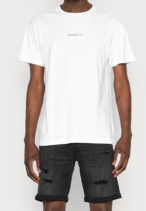 T-shirt en coton blanc avec un col rond et des manches courtes, arborant un petit logo noir sur le devant. Porté avec un short en denim noir déchiré.