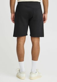 Shorts en coton noir avec une taille à cordon, une poche arrière et une coupe décontractée, se terminant juste au-dessus du genou. Texture lisse, détails minimaux.