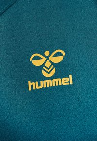 Gult stiliseret bi-logo og tekst "hummel" trykt på teal tekstureret stof, sandsynligvis sportstøj materiale.