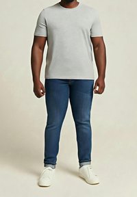 Homme portant un t-shirt gris clair à manches courtes, un jean bleu foncé retroussé au niveau des chevilles, et des baskets blanches, debout devant un fond uni.
