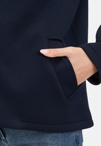 Granatowy sweatshirt o gładkiej fakturze, z boczną kieszenią i zaokrąglonym dołem, wykończony starannie wszytym szwem.