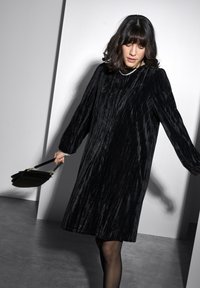 Robe en velours noir à manches longues et coupe ample. Caractérisée par des lignes de texture verticales et un col rond. Sac à main noir tenu dans une main.