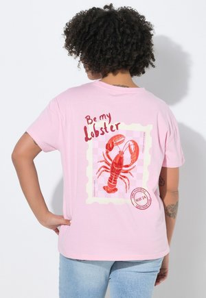 Person med krøllet hår iført en pink T-shirt med en rød hummer, teksten "Be my Lobster" og et cirkulært stempelmotiv på ryggen.