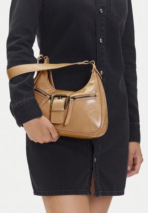 Person in schwarzem Kleid hält eine beigefarbene Lederhandtasche mit vorderem Schnallenverschluß und Reißverschlusstasche, die am Unterarm hängt.