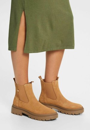 Bottines en daim camel à semelles épaisses portées avec une jupe verte fendue sur le côté, visibles sur des jambes nues sur un fond neutre.