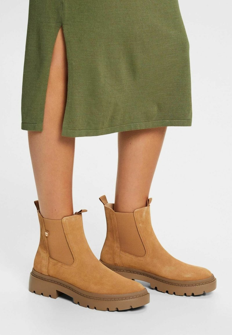 Bottines en daim camel à semelles épaisses portées avec une jupe verte fendue sur le côté, visibles sur des jambes nues sur un fond neutre.