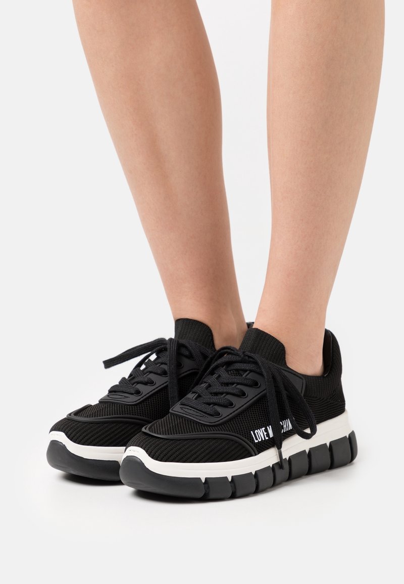 Baskets en maille noire avec des semelles épaisses segmentées blanches et noires portées sur des jambes nues, avec le texte "LOVE MOSCHINO" sur le côté.
