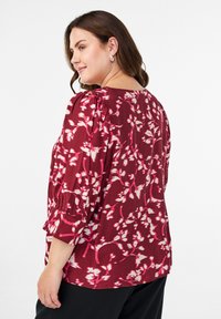 Burgundy-blouse har hvit blomsterpryd, med puffede ermer og en litt løs passform. Glatt tekstur med rund hals.