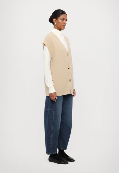 Beige strikket ærmeløs vest med V-hals, udstyret med fire knapper. Kombinationen er med løse blå denim bukser og sorte flade sko.