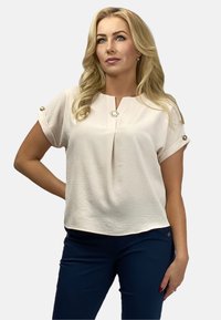 Jasnoróżowa bluzka z krótkim rękawem, okrągłym dekoltem, fałdą z przodu i ozdobnymi guzikami na rękawach, wykonana z miękkiego, teksturowanego materiału.