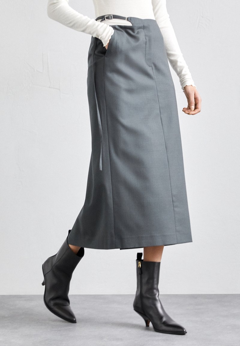 Osoba ubrana w szarą spódnicę midi z wysokim stanem i bocznymi kieszeniami oraz czarne botki z noskiem w szpic, w trakcie chodzenia na tle jednolitego tła.