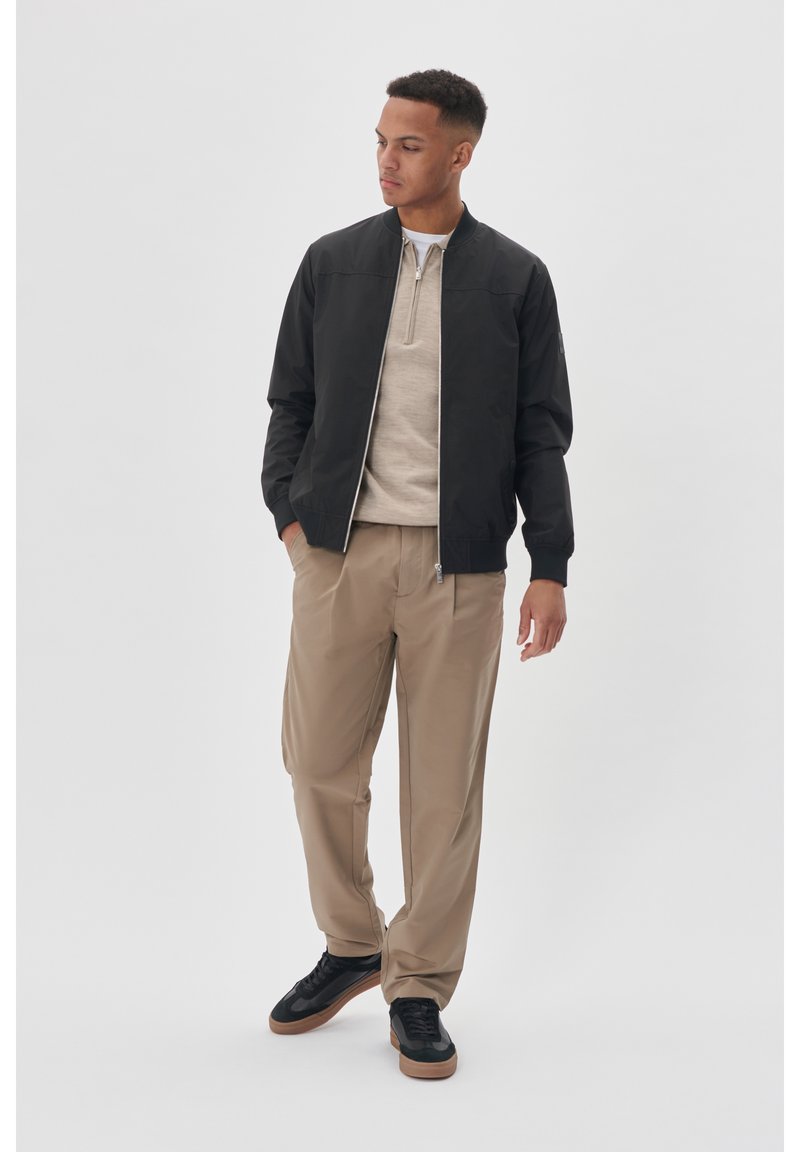 Crna bomber jakna s patentnim zatvaračem sprijeda, bež lagana veste ispod, bež hlače i crne tenisice. Glatka tekstura i elegantan dizajn.
