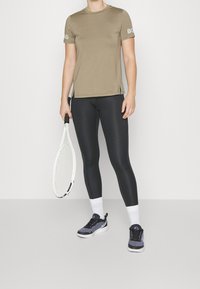 Camisa atlética de manga corta en un tono verde oliva claro con aberturas laterales, combinada con leggings negros y zapatillas de deporte en blanco y negro, sosteniendo una raqueta de tenis.