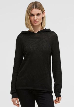 Woman with straight blonde hair wearing a black hooded sweater with subtle textured text standing against a plain light background.