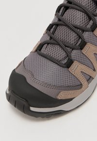 Chaussure de randonnée grise avec tige en mesh, capuchon en caoutchouc noir, accents beiges et détails réfléchissants argentés. Dispose d'un système de laçage texturé.
