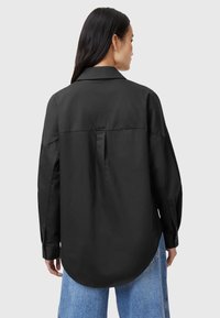 Maglietta nera con colletto classico, vestibilità over size, polsini con bottone e orlo arrotondato. Realizzata in tessuto liscio con un dettaglio di piega sul retro.