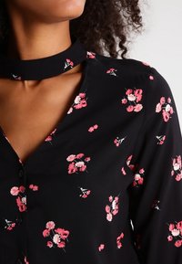 Black floral blouse with a V-neck and choker collar, featuring small pink and white flowers, button closure, and soft fabric texture.