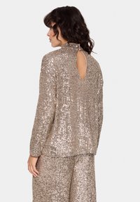 Top a maniche lunghe in paillettes dorate-beige lucide, con colletto alto e schiena scoperta. Vestibilità comoda e texture brillante con linee verticali.