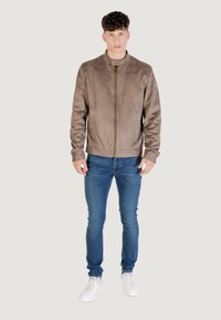 Giubbotto in finta pelle marrone con colletto alto, chiusura con zip e texture liscia, abbinato a jeans blue e sneakers bianche.