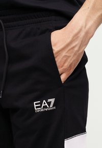 Χέρι στην τσέπη των μαύρων παντελονιών Emporio Armani EA7 με κορδόνι στη μέση και λευκή λεπτομέρεια κοντά στο γόνατο.