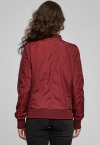 Veste bomber bordeaux avec une texture lisse et brillante, poignets et taille côtelés, ornée de fronces le long du dos et des manches.