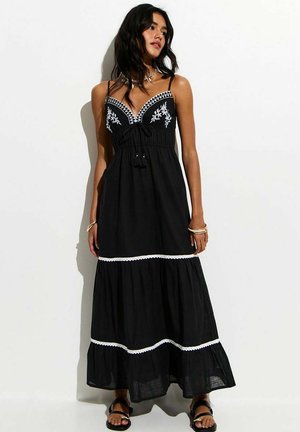 Kobieta w czarnej sukni maxi z białym kwiatowym haftem i koronkową lamówką, stojąca w zrelaksowanej pozie na prostym białym tle.