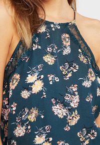 Blommig halterneck-topp i mörk teal-fabric som har flerfärgade blommor och spetsdetaljer kring halsringningen, med avslappnad passform och öppen rygg.