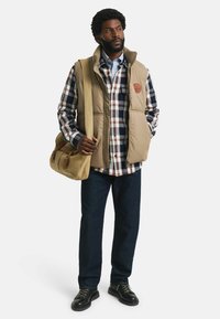 Chaleco acolchado beige sobre una camisa de cuadros en azul, rojo y blanco. Jeans oscuros y zapatos negros. Bolsa de hombro marrón hecha de lona.