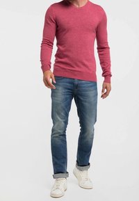 Pull rouge à col en V, ajusté, associé à un jean bleu clair et des baskets blanches. Le pull a des poignets et un ourlet côtelés.