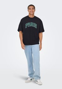 Zwart oversized T-shirt met "PARIS" in groene boogletters. Gecombineerd met lichtblauwe spijkerbroek en witte sneakers met groene accenten.