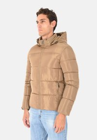 Giacca puffer beige con zip frontale, cappuccio e tasche laterali. Realizzata in tessuto lucido e imbottito con motivi di cucitura orizzontale.