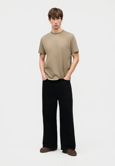 Νέος άνδρας που στέκεται με τα χέρια στις τσέπες, φοράει μπεζ κοντομάνικο πουκάμισο, φαρδιά μαύρα παντελόνια και καφέ παπούτσια σε απλό ανοιχτό φόντο.