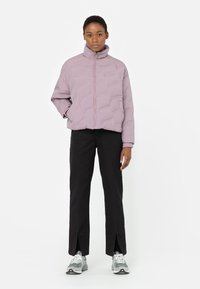 Chaqueta de plumas lila con cuello alto, que presenta un patrón acolchado. Combinada con pantalones negros y zapatillas grises.