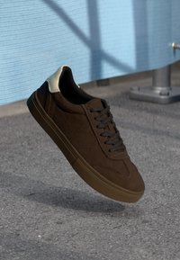 Brown suede sneakers with a beige leather accent at the heel. features a low profile, flat rubber sole, and dark brown laces.