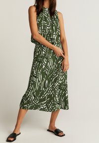 Grünes ärmelloses Kleid mit weißen abstrakten Mustern, tailliertem Bund und fließendem Rock. Getragen mit schwarzen flachen Slidesandalen.
