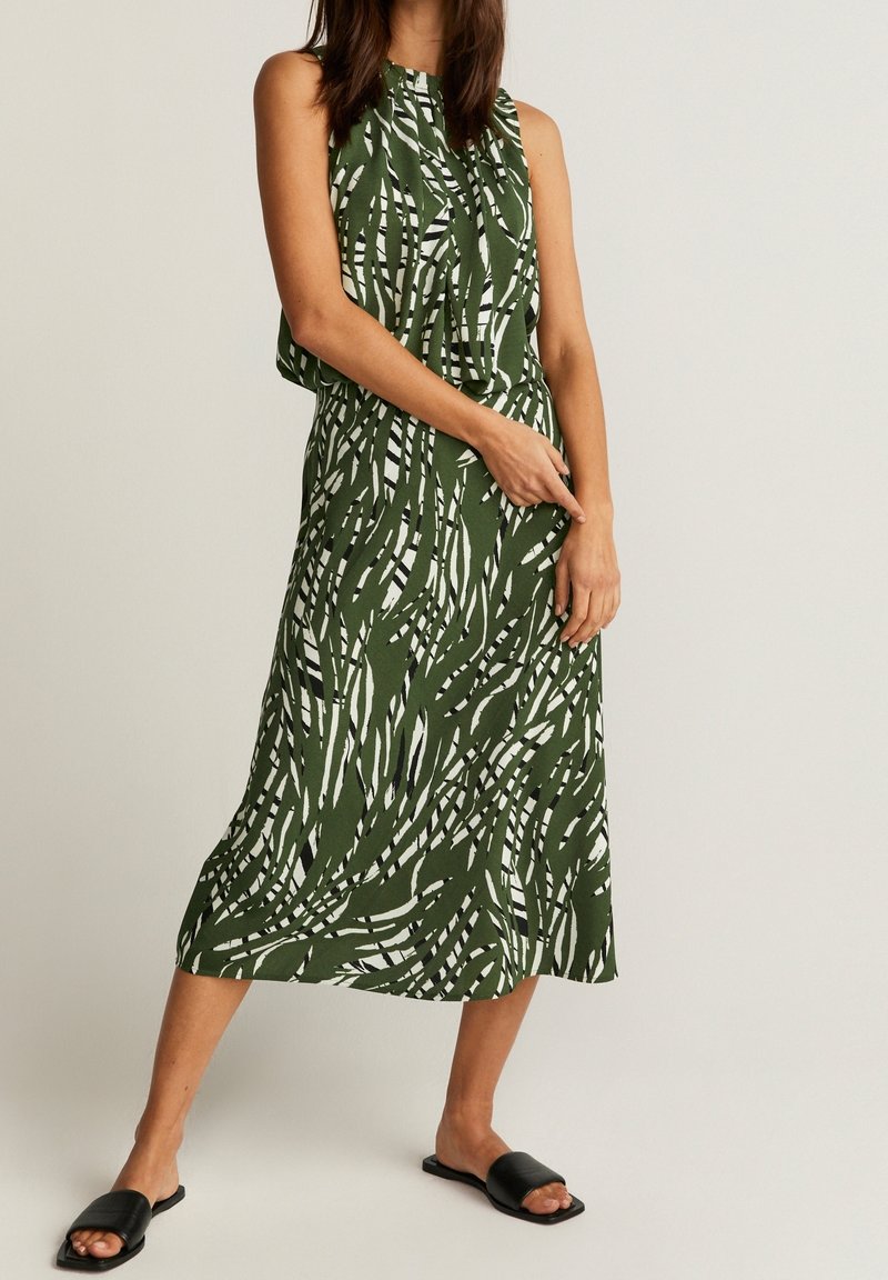 Grünes ärmelloses Kleid mit weißen abstrakten Mustern, tailliertem Bund und fließendem Rock. Getragen mit schwarzen flachen Slidesandalen.