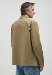 Un homme aux cheveux mi-longs porte une veste beige et un jean bleu, se tenant de profil contre un fond clair.