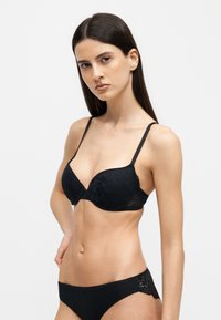 Femme aux longs cheveux foncés porte un soutien-gorge en dentelle noire et des sous-vêtements noirs assortis, posant avec une main sur la hanche devant un fond uni.