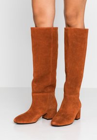 Botas altas de ante marrón con punta cuadrada y tacón ancho. Textura suave, diseño minimalista sin hardware visible ni patrones.