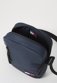 Navy blue crossbody bag made of smooth synthetic material, featuring a zippered closure, adjustable strap, and logo patch on the front.