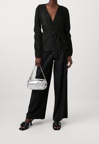 Blouse noire plissée portefeuille à manches longues, taille cintrée, associée à un pantalon noir taille haute et un sac à main métallique argenté. Sandales plates noires.