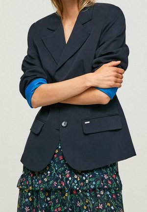 Vrouw met een donker colbert met opgestroopte blauwe mouwen over een bloemrijke, gerimpelde jurk, met haar armen gekruist over haar borst.