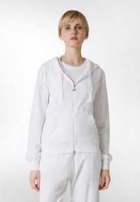 Weiße Zip-Hoodie aus weichem Stoff, mit Kapuze, Fronttaschen, gerippten Bündchen und einem glatten Reißverschluss. Über einem weißen Shirt getragen.