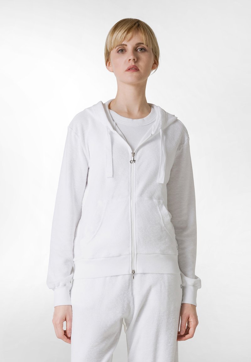 Weiße Zip-Hoodie aus weichem Stoff, mit Kapuze, Fronttaschen, gerippten Bündchen und einem glatten Reißverschluss. Über einem weißen Shirt getragen.