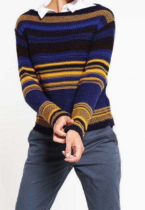 Persona che indossa un maglione a righe blu navy, blu, nero e giallo sopra una camicia bianca con colletto e pantaloni grigi, mentre sistema le maniche.
